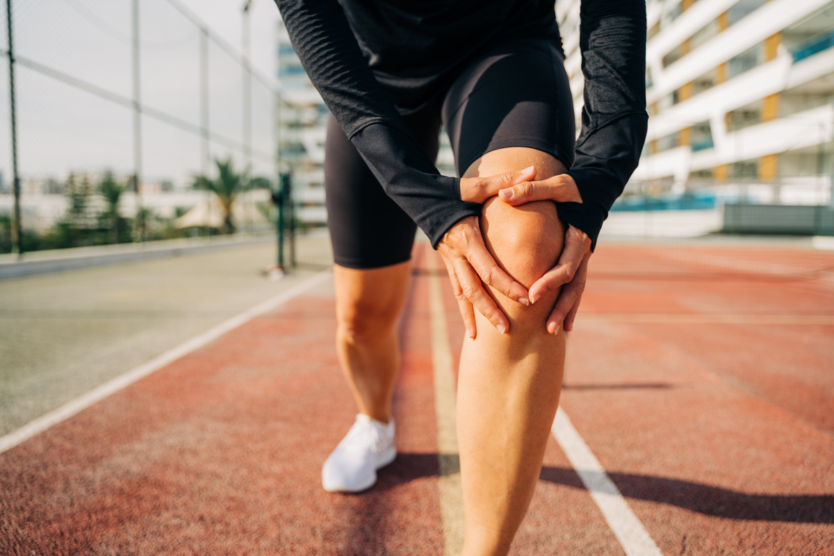 Close up of sport woman holding knee with hands in pain after suffering injury running. Runner woman sit on road with knee injury and pain